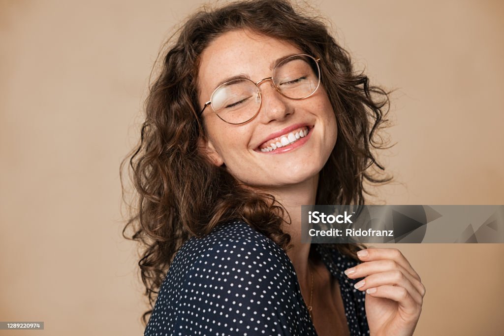 Joyful natural young woman smiling Casual cheerful woman with eyeglasses smiling at camera on cream background. Close up of happy young woman laughing with eyeglasses. Beautiful natural girl having fun with closed eyes showing a big grin. Women Stock Photo