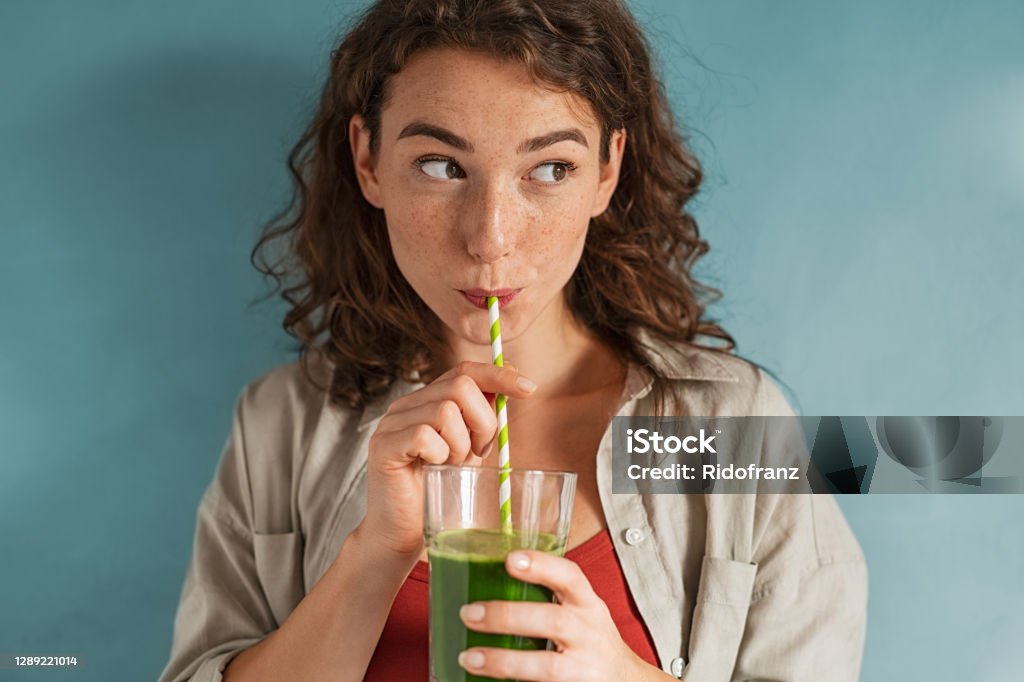 Young woman drinking detox juice with straw on blue wall Beautiful woman drinking an organic green smoothie. Fit young woman drinking detox juice using paper straw isolated against blue background. Healthy girl enjoy detox drink and looking away. Healthy Eating Stock Photo