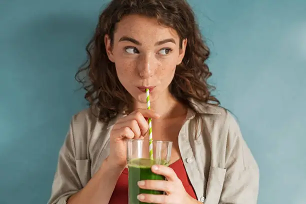 Photo of Young woman drinking detox juice with straw on blue wall