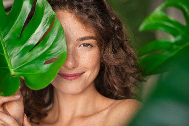 Beauty natural woman covering her face with tropical leaf Close up face of beautiful young woman covering her face by green monstera leaf while looking at camera. Natural smiling girl with green palm leaf. Portrait of beauty woman with natural makeup and freckles on skin standing behind big green leaves. purity stock pictures, royalty-free photos & images