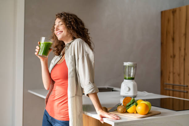 Woman drinking detox drink at home Happy young woman standing in kitchen and holding glass of detox juice. Cheerful girl drinking healthy smoothie at home. Beautiful smiling woman drinking green vegetable smoothie. smoothie stock pictures, royalty-free photos & images