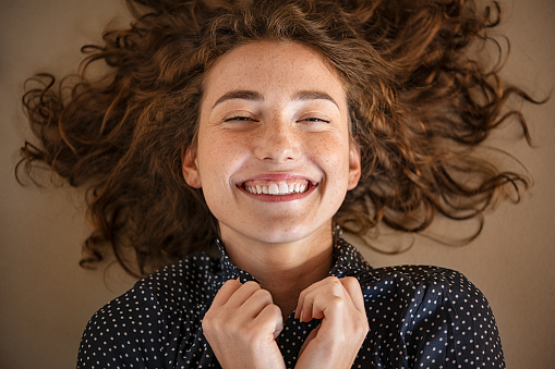 Portrait of laughing young woman with closed eyes smiling in joy. Close up face of cheerful girl looking at camera while lying on floor. Happy girl laughing while looking at camera and lying on floor.