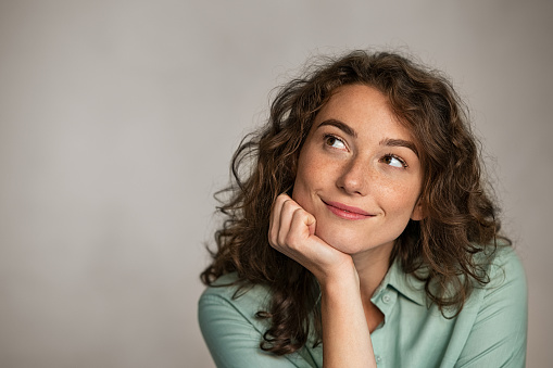 Portrait of young thoughtful woman with hand on chin having an idea against grey background. Beautiful pensive woman looking away while thinking. Close up face of natural girl planning her future isolated on gray wall with copy space.