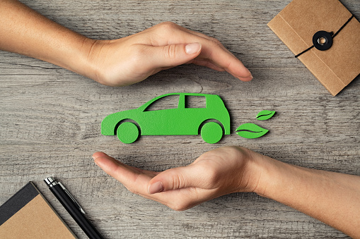 Close up of hands of young woman protecting icon of green eco-friendly car over wooden table. Top view of woman hands protecting electric car with care, mobility. Car insurance and green automotive business concept.
