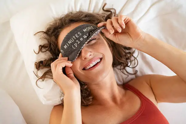 Top view of tired young woman sleeping on bed. Close up face of beautiful sleepy girl wearing funny eye mask while peeping with one eye. High angle view of woman covering her eyes with sleeping mask with five minutes more text.