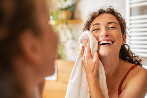 Young woman wiping her face with white towel after waking up in the morning. Portrait of beautiful happy smiling girl holding clean towel near facial skin after washing face. Happy woman cleaning and drying skin with napkin.