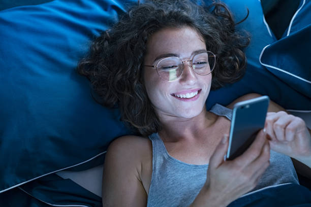 Happy smiling woman using smartphone late in night Relaxed young woman using smartphone and lying on bed while wearing spectacles at night. Happy girl wearing big eyeglasses and messaging with cellphone late at night. Close up face of smiling carefree girl using social network on mobile phone before sleep. relaxation lying on back women enjoyment stock pictures, royalty-free photos & images