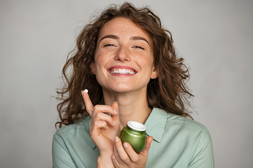 Attractive young woman applying cream on her face full of freckles. Natural girl holding biological and vegan moisturizer jar while looking at camera. Portrait of beauty casual woman with daily lotion for soft hydrated skin isolated on grey background.