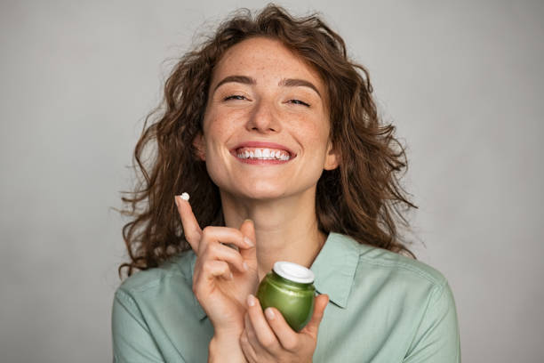 Beautiful smiling woman applying facial cream from green jar Attractive young woman applying cream on her face full of freckles. Natural girl holding biological and vegan moisturizer jar while looking at camera. Portrait of beauty casual woman with daily lotion for soft hydrated skin isolated on grey background. fresh faced stock pictures, royalty-free photos & images