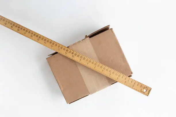 Cardboard shipping box placed on white background. Leaning on it is a wooden yardstick whit centimeters and inches fractions scales. Association of measuring a box for delivery.