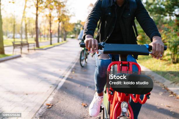 Photographic Closeup Of A Student Boys Body With A Backpack On His Back Carrying A Red Shared Electric Bike Along A Treelined Park Path At Sunset Stock Photo - Download Image Now