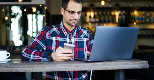 istock Cheerful male blogger in eyeglasses using computer and cellphone in cafe 1289226526