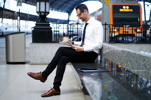 Side view of full body content male manager in formal wear resting on bench while waiting for train with newspaper