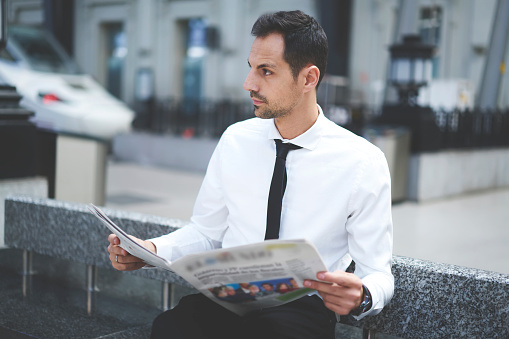 Concentrated male entrepreneur in formal outfit sitting on bench and looking away while reading newspaper and thinking about business news