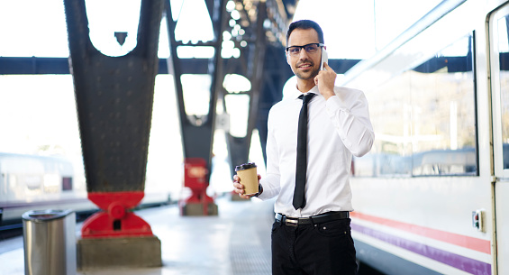 Confident male manager in elegant formal clothes and spectacles standing near train on railway station and talking on smartphone while having cup of coffee