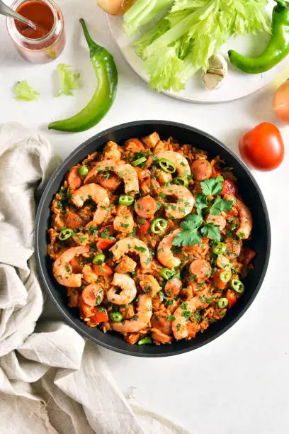 Creole jambalaya with chicken, smoked sausages and vegetables in frying pan on white stone background. Top view, flat lay, close up
