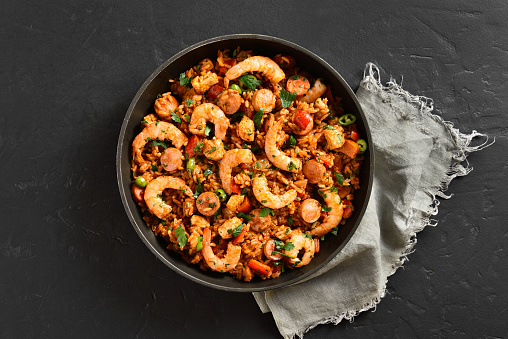 Creole style jambalaya with chicken, smoked sausages and vegetables in frying pan over black stone background. Top view, flat lay, close up