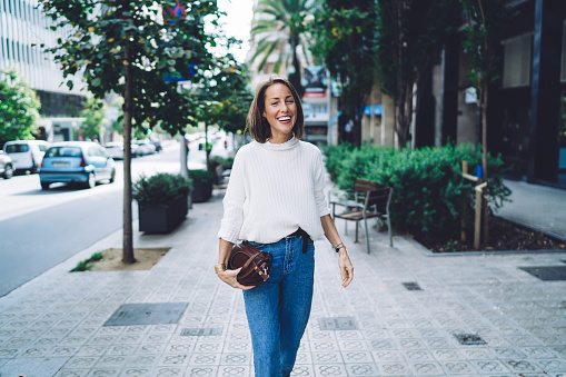 Happy female in casual outfit with brown leather handbag laughing happily while walking on city street with modern buildings and greenery