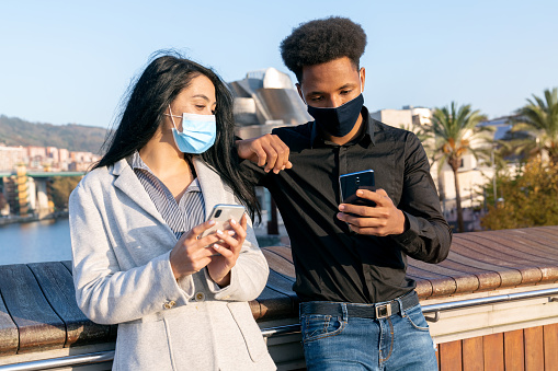 Portrait of a young couple on the street using their mobile phone writing text with a face mask due to the 2020 covid-19 coronavirus pandemic boy witn afro style hair