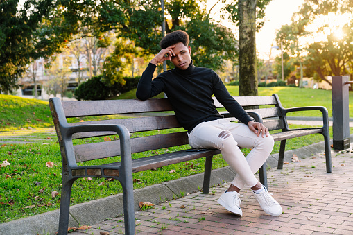 Fashionable portrait of a handsome Moroccan man with afro hair sitting on a park bench in urban style clothes on a beautiful sunset