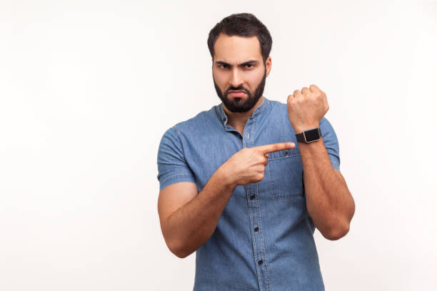 hombre puntual molesto con barba con camisa azul apuntando con el dedo al reloj de pulsera maqueta pantalla y expresando insatisfacción con la hora tardía, la fecha límite - impaciencia fotografías e imágenes de stock