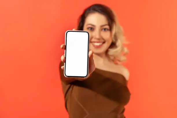 Positive smiling woman holding and showing smartphone with empty white display, freespace for your advertisement, tariffs and bonuses, mobile app. Indoor studio shot isolated on orange background