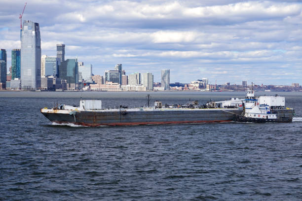 A tug boat helps a commercial shipping barge navigate the waters of New York Harbor with the Jersey City skyline in the background
