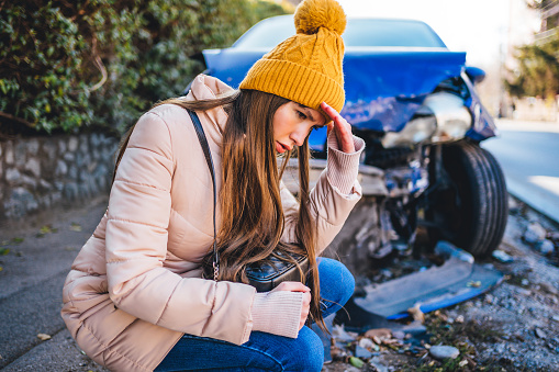 Young frustrated woman crouches next to wrecked car after a car accident