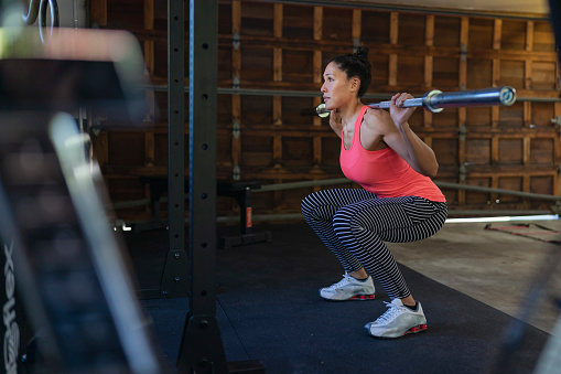 A fit Pacific Islander woman does squats with a barbell while doing a cross training workout in the home gym set up in her garage. The woman is staying healthy while isolating at home and social distancing during the Covid-19 pandemic.
