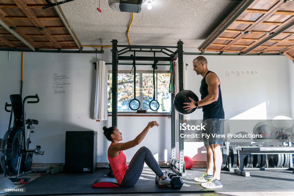 Woman exercising with personal trainer in home gym Beautiful and fit Pacific Islander woman and muscular Caucasian man doing an abdominal strength workout with a weighted ball in a home gym set up in the garage of their home. Gym Stock Photo
