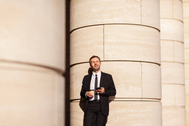 istock Middle aged buisnessman looking away dreamily and squinting in sun leaning on column with takeaway coffee cup and cellphone in his hands 1289249993
