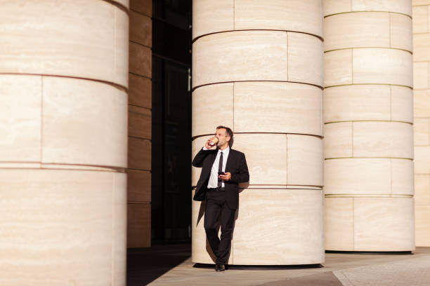 istock Wide shot of middle aged buisnessman drinking takeaway coffee while leaning on column outside with cell phone in his hand 1289250001