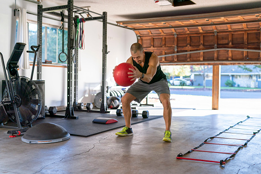 A athletic man uses a medicine ball while doing a cross training workout in the gym he has set up in the garage of his home. He is staying healthy and active while isolating at home during the Covid-10 pandemic.