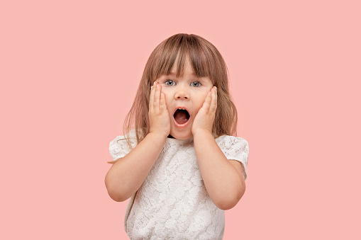 Human emotion of surprise on the face of a little blonde girl in a white t-shirt. Pink isolated background.