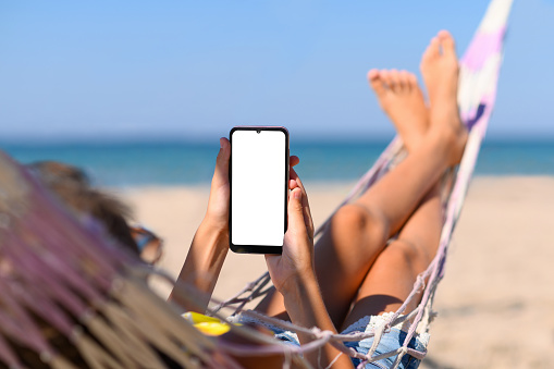 Smartphone with a blank white display in the hands of a girl against the background of the sea. The girl lies on a hammock on the beach with a phone in her hands.