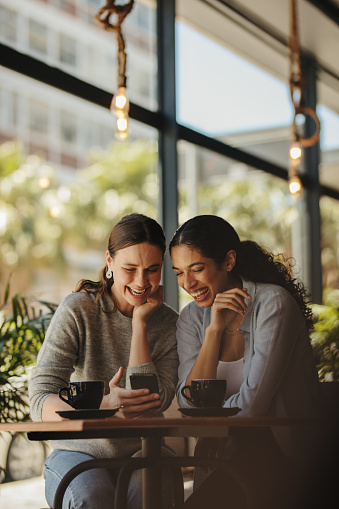 Friends sitting in a cafe looking at a smartphone and smiling. Two women meeting in a coffee using a mobile phone.