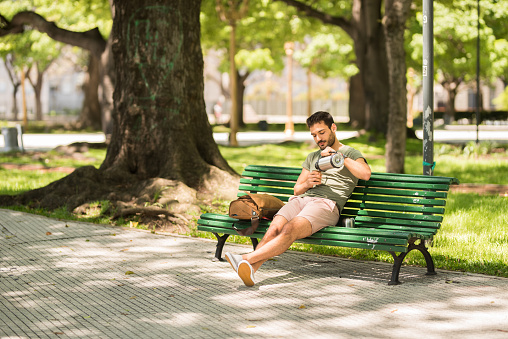 Young man pouring himself a cup of mate tea from a flask while relaxing on a park bench in the summer