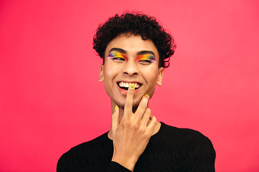 Happy young man wearing rainbow colored eye shadow and smile face nail paint. Gay male smiling with hand on face against red background.