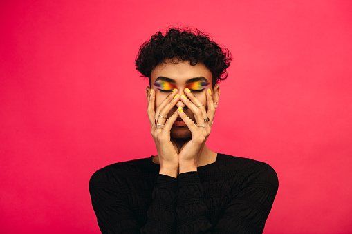 Gay man wearing rainbow colored eye shadow. Androgynous male holding his face with eyes closed on red background.