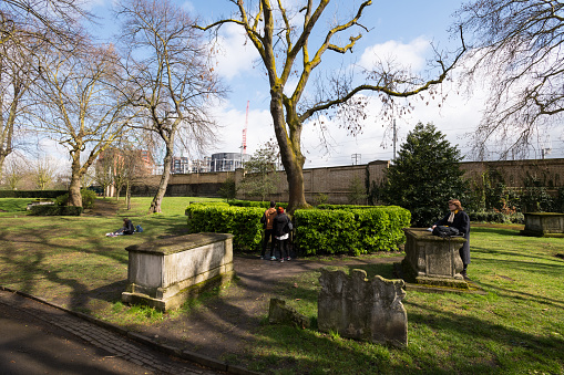 London, UK - Mar 19, 2019: People visiting the Graves stacked at the base of a tree at St Pancras Old Church in Somers Town late in the day.