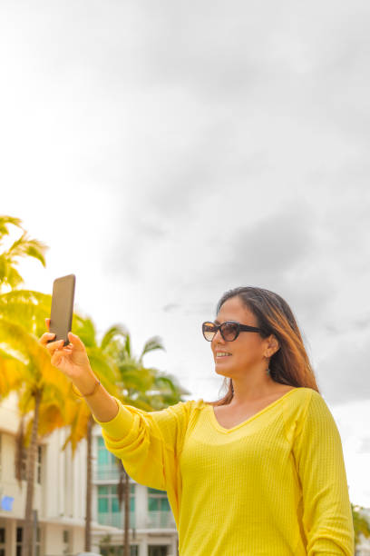 Smiling young woman taking a photography by her smart phone in Miami Beach, Florida, USA Young latin woman taking a photography by her cellphone in Miami Beach, Florida, USA. She's wearing a nice fashionable black eyewear, a yellow shirt, a red purse and jeans, in a cloudy day in summer season, on back the Miami Art Deco District. 1 hotel south beach stock pictures, royalty-free photos & images