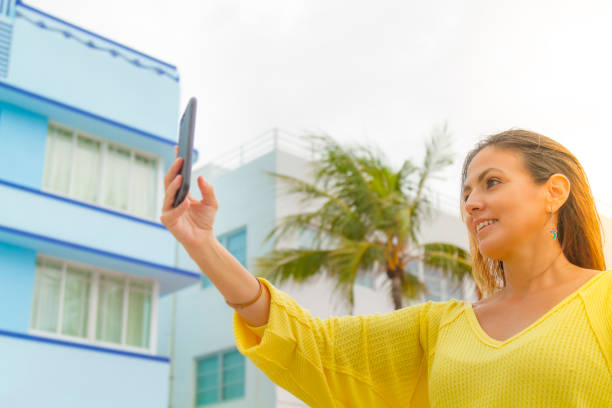 Smiling young woman taking a photography by her smart phone in Miami Beach, Florida, USA Young latin woman taking a photography by her cellphone in Miami Beach, Florida, USA. She's wearing a nice fashionable black eyewear, a yellow shirt, a red purse and jeans, in a cloudy day in summer season, on back the Miami Art Deco District. 1 hotel south beach stock pictures, royalty-free photos & images