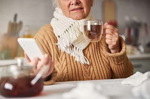 Cropped photo of tired aging lady with scarf around neck drinking tea and looking at smartphone screen