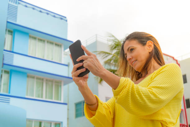 Smiling young woman taking a photography by her smart phone in Miami Beach, Florida, USA Young latin woman taking a photography by her cellphone in Miami Beach, Florida, USA. She's wearing a nice fashionable black eyewear, a yellow shirt, a red purse and jeans, in a cloudy day in summer season, on back the Miami Art Deco District. 1 hotel south beach stock pictures, royalty-free photos & images