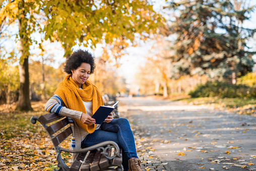 Mid adult African American woman sitting on bench and reading a book on autumn day in the park.