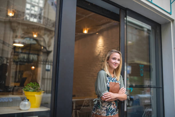 Welcoming Blonde Cafe Owner Leaning Against Front Door Relaxed Argentine female cafe owner in late 20s standing at open front door of cafe in apron with arms crossed. small business saturday stock pictures, royalty-free photos & images