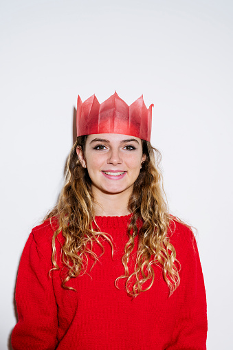 Partial front view portrait of 14 year British old girl with long curly hair in red Christmas cracker hat and casual sweater smiling at camera against white background.