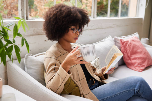istock Young woman drinking coffee and reading a book on her sofa 1289318340