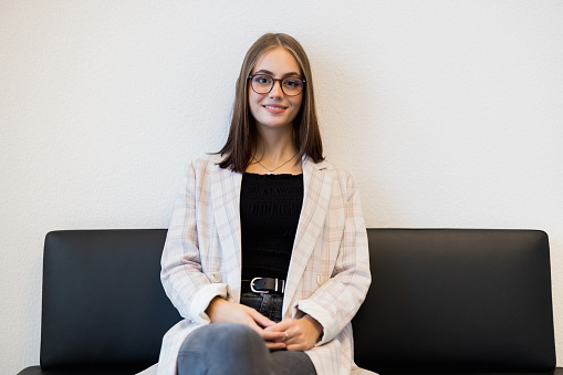 Happy smiling female business entrepreneur waering glasses in casual businesswear sitting on black leather bench. Reduced modern Businesswoman Portrait.