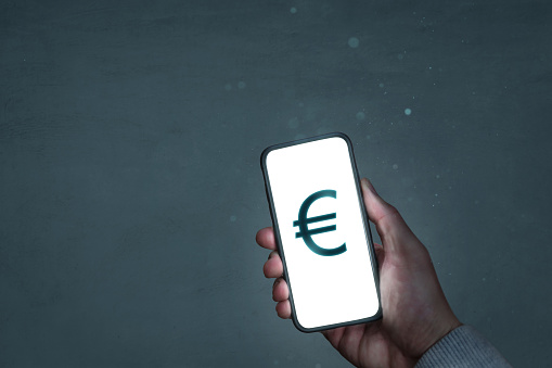 A modern smartphone held in one hand. The screen glows and shows a Euro currency symbol.